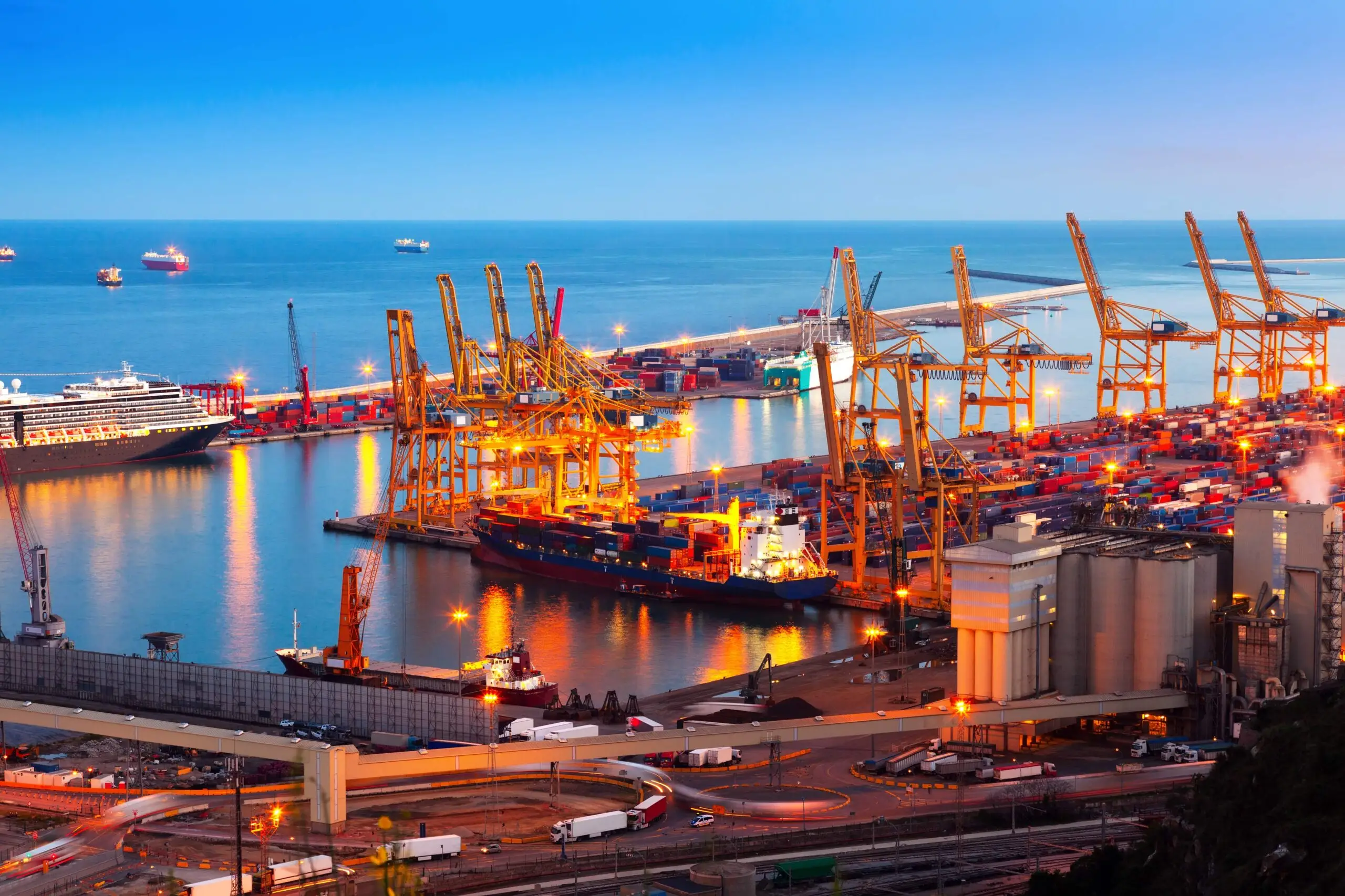Puerto industrial de noche con grúas pórtico, buques atracados y camiones en movimiento